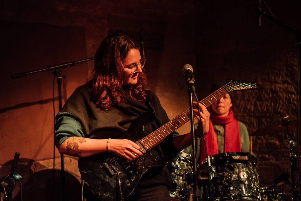 Frau mit dunklen Haaren steht im schummrigen Licht auf einer kleinen Bühne und spielt E-Gitarre