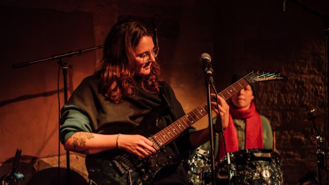 Frau mit dunklen Haaren steht im schummrigen Licht auf einer kleinen Bühne und spielt E-Gitarre