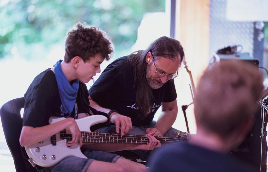 Auf dem Bild sind zwei Personen zu sehen. Die eine Person ist jung, hält eine E-Gitarre und hat die eine Hände um den Gitarrenhals platziert. Die andere Person sittzt daneben, beugt sich über die Gitarre und hilft beim spielen.