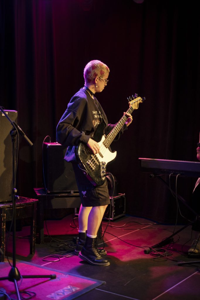 Performance bei einer Jam Session. Eine Person spielt auf der Bühne Bassgitarre und im Hintergrund werden weitere Instrumente und die Bühnenbeleuchtung dargestellt