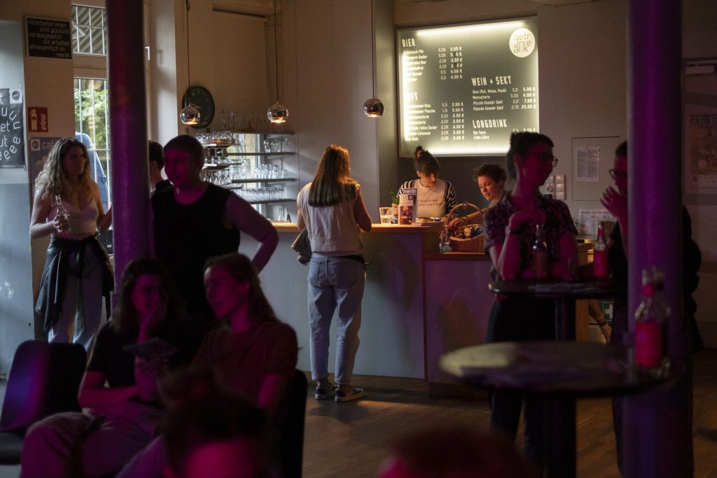 Es wird ein Raum gezeigt, in dem sich viele Menschen aufhalten. Es gibt eine Bar an der eine Frau sich etwas kauft. Menschen unterhalten sich und feiern einen schönen Abend.